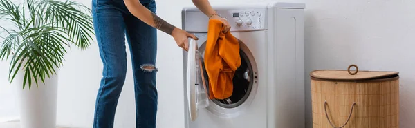 Vista parcial de la mujer poniendo la ropa en la lavadora cerca de la cesta de la ropa y la planta, pancarta - foto de stock