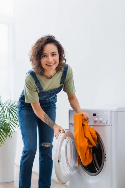 Alegre dona de casa olhando para a câmera enquanto segurando roupas perto da máquina de lavar roupa — Fotografia de Stock