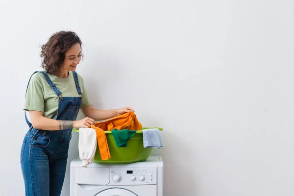 Fröhliche Frau hält Kleidung in der Nähe von Waschschüssel auf Waschmaschine — Stockfoto