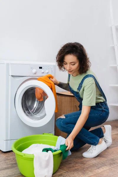 Brünette Frau zieht Kleidung aus Wäschekorb und steckt sie in Waschmaschine — Stockfoto