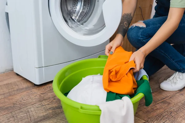 Vue recadrée d'une femme tatouée tenant des vêtements près de la laveuse et du bol à linge — Photo de stock