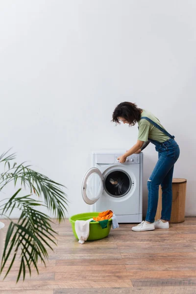 Brunette woman operating washing machine near clothes in laundry bowl — Stock Photo