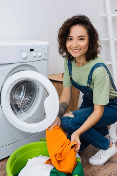 Brunette woman smiling at camera while holding laundry near washing machine — Stock Photo