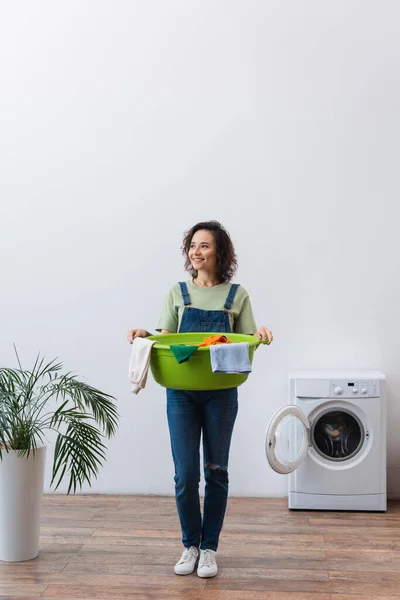 Full length view of happy housewife holding laundry bowl with clothes near plant and washing machine — Stock Photo