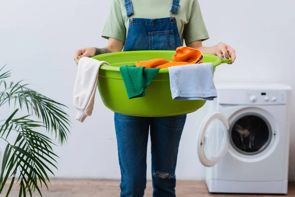 Обрезанный вид на домохозяйку, держащую миску для стирки возле размытой стиральной машины в ванной комнате — стоковое фото