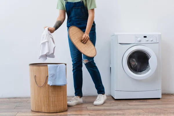 Vue partielle de la femme en salopette denim tenant des vêtements près du panier à linge et de la machine à laver — Photo de stock