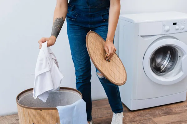 Vue recadrée d'une femme tatouée tenant des vêtements près du panier à linge et de la rondelle — Photo de stock