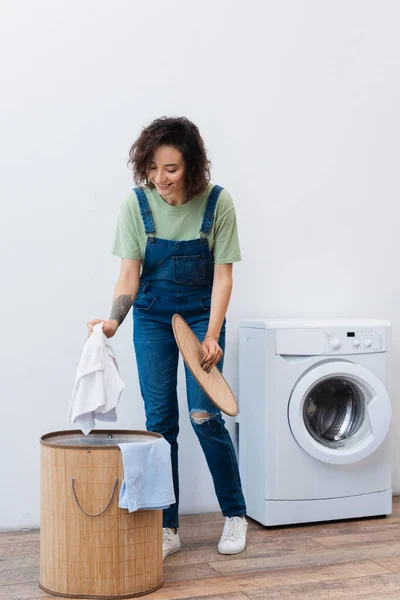 Mulher sorrindo segurando roupa perto da cesta e máquina de lavar roupa — Fotografia de Stock