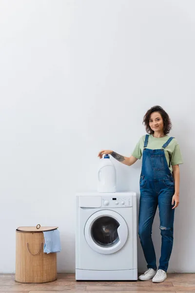 Lächelnde Hausfrau blickt in die Kamera neben Waschmittelflasche auf Waschmaschine — Stockfoto