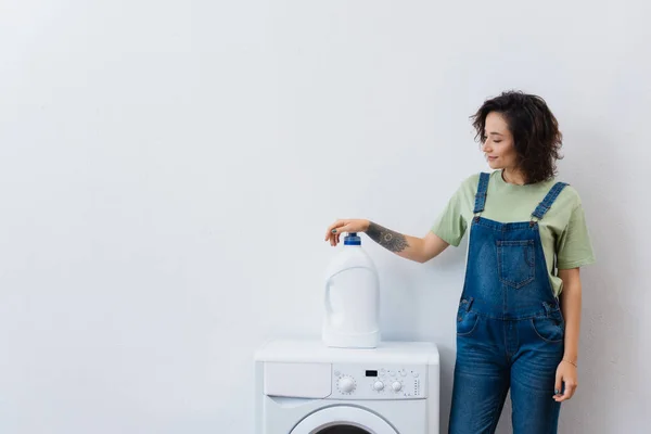 Усміхнена домогосподарка стоїть біля пляшки миючого засобу на пральній машині — стокове фото