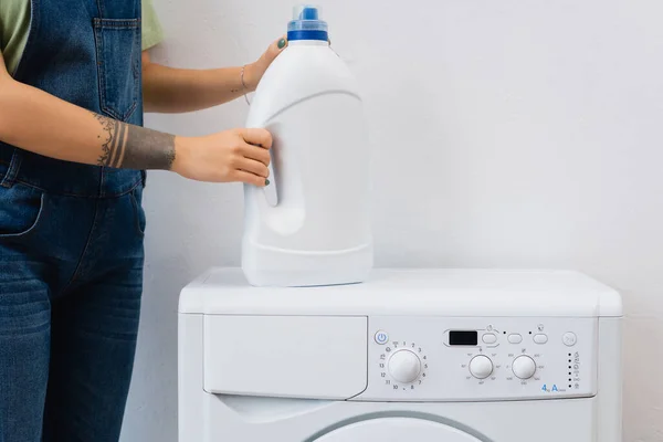 Частковий вид татуйованої жінки, що тримає пляшку миючого засобу біля пральної машини — стокове фото