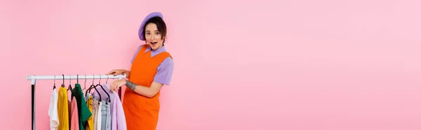 Amazed woman in purple beret looking at camera near rack with clothes isolated on pink, banner — Stock Photo