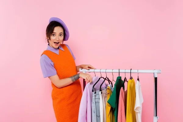 Astonished woman in orange sleeveless dress looking at camera near rack with clothes isolated on pink — Stock Photo