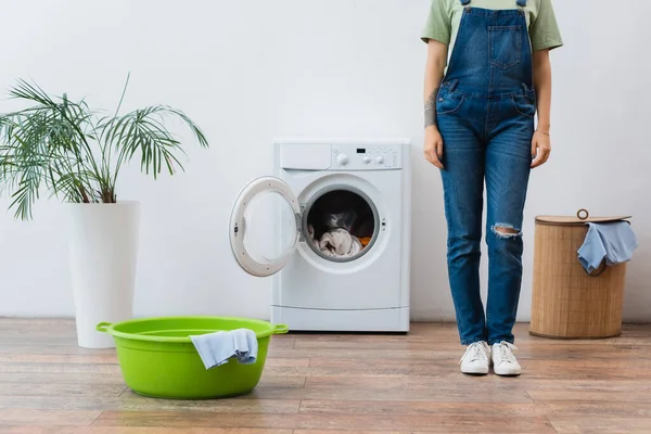 Cropped view of woman in denim overalls standing near washing machine, laundry bowl and basket — Stock Photo