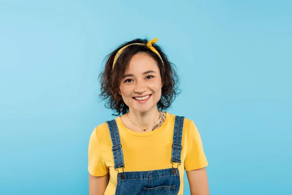 Pretty woman in headband and denim overalls smiling at camera isolated on blue — Stock Photo