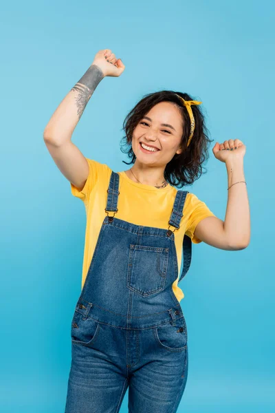 Aufgeregte Frau in Jeans-Overalls zeigt Siegergeste, während sie isoliert auf blau in die Kamera blickt — Stockfoto