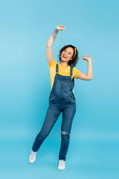 Полный вид взволнованной женщины в джинсовом комбинезоне, показывающей жест триумфа на голубом — стоковое фото