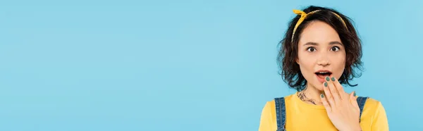 Astonished woman covering open mouth with hand while looking at camera isolated on blue, banner — Stock Photo