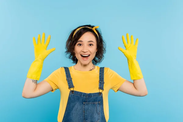 Femme au foyer gaie en gants en caoutchouc jaune regardant la caméra isolée sur bleu — Photo de stock