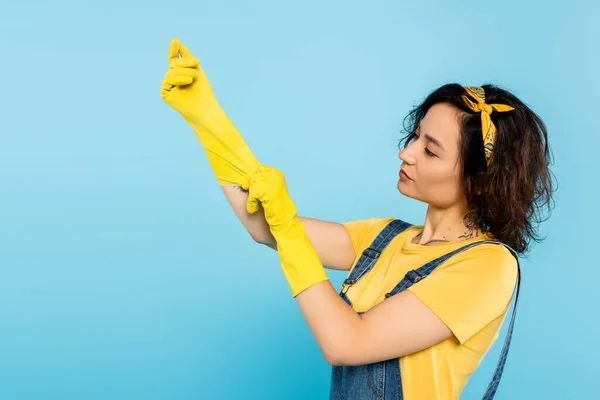 Morena mujer poniéndose guantes de goma amarillos aislados en azul - foto de stock