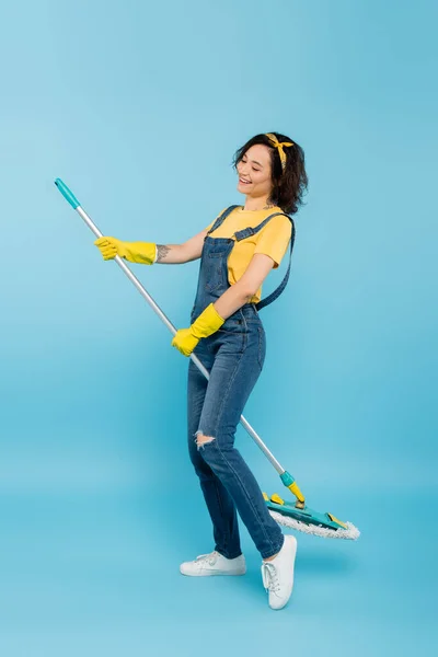 Щаслива домогосподарка в жовтих гумових рукавичках і джинсовому комбінезоні позує з мопедом на синьому — стокове фото