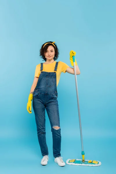 Vue pleine longueur de la femme en salopette en denim et gants en caoutchouc posant avec une serpillière sur bleu — Photo de stock