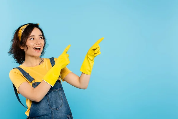 Mujer excitada en guantes de goma amarillos mirando hacia otro lado y señalando con los dedos aislados en azul - foto de stock