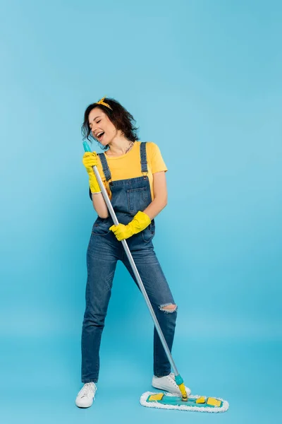 Cheerful housewife in denim overalls and rubber gloves singing with mop on blue — Stock Photo