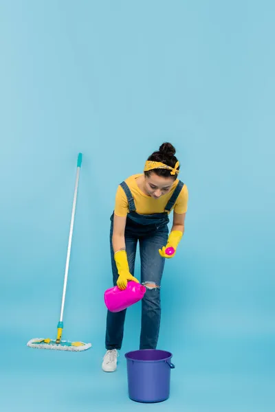 Femme au foyer en salopette denim et gants en caoutchouc versant du détergent dans le seau près de la serpillière sur bleu — Photo de stock