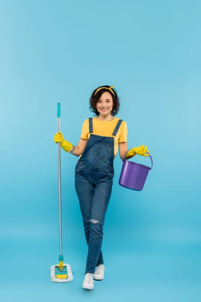 Vista completa de la mujer en guantes de goma amarilla posando con cubo y fregona en azul - foto de stock