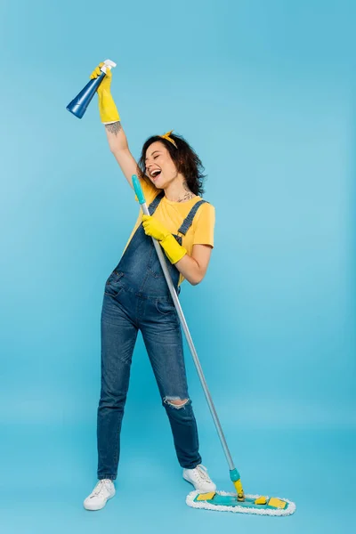 Excited woman in rubber gloves and denim overalls singing with detergent and mop on blue — Stock Photo