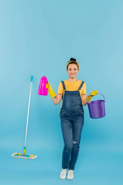 Sorridente dona de casa em denim macacão segurando detergente e balde perto esfregona em azul — Fotografia de Stock