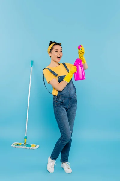 Mujer excitada en overoles de mezclilla sosteniendo detergente mientras está de pie cerca de fregona en azul - foto de stock