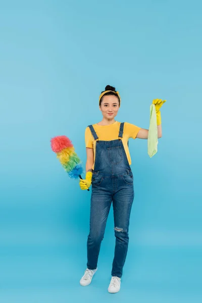 Vue pleine longueur de la femme au foyer avec brosse à poussière colorée et chiffon regardant la caméra sur bleu — Photo de stock