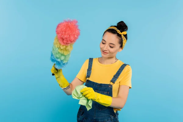 Femme souriante en salopette denim tenant chiffon et brosse à poussière colorée isolé sur bleu — Photo de stock