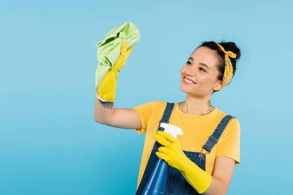 Mujer feliz en guantes de goma amarillos sosteniendo trapo y botella de aerosol con limpiador aislado en azul - foto de stock