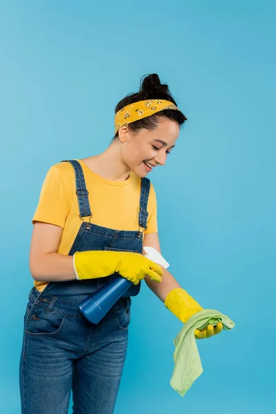 Sonriente ama de casa rociando detergente en trapo aislado en azul - foto de stock