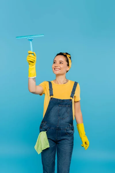 Happy housewife with rag in pocket of denim overalls holding window cleaner isolated on blue — Stock Photo