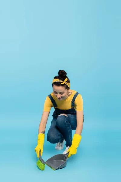 Positive Hausfrau in Jeans-Overalls und Gummihandschuhen fegt Boden auf blau — Stockfoto