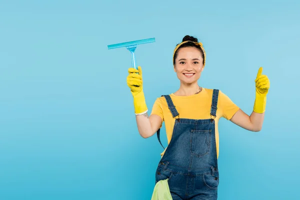 Joyful housewife with window cleaner showing thumb up while looking at camera isolated on blue — Stock Photo