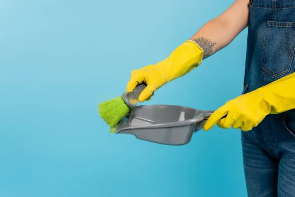 Partial view of tattooed housewife in yellow rubber gloves holding scoop and broom isolated on blue — Stock Photo