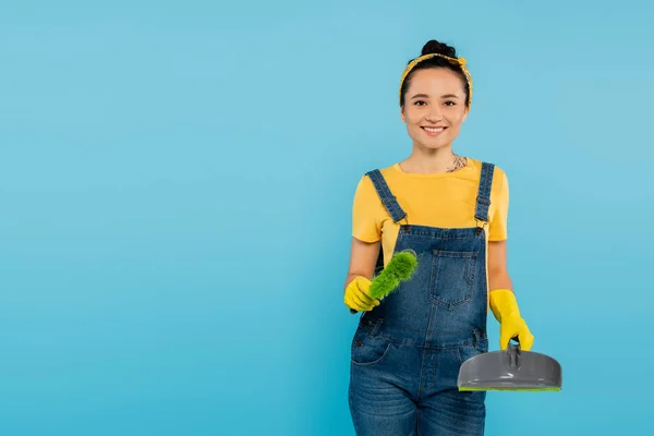 Alegre ama de casa con cucharada y escoba sonriendo a la cámara aislado en azul - foto de stock