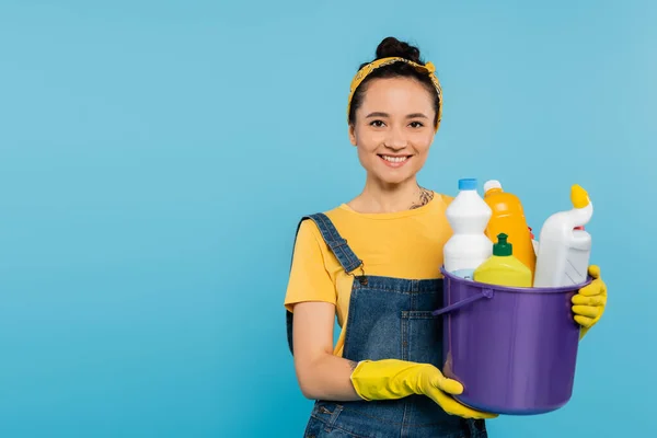 Jovem dona de casa em luvas de borracha amarela segurando balde com detergentes isolados em azul — Fotografia de Stock