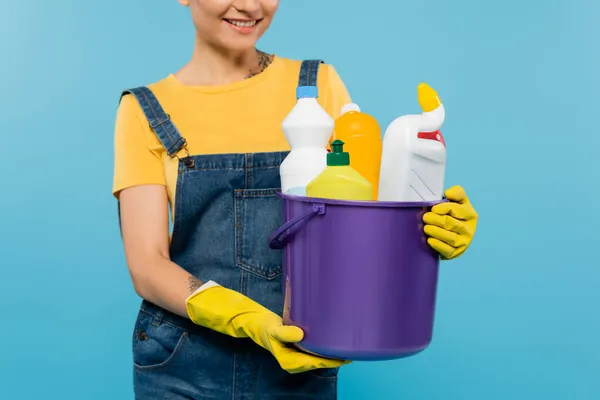 Vista recortada de ama de casa sonriente en guantes de goma amarilla sosteniendo cubo con limpiadores aislados en azul - foto de stock