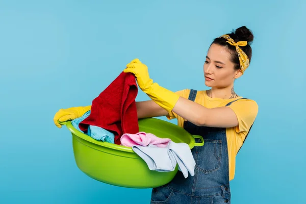 Housewife in yellow rubber gloves looking at clothing in laundry bowl isolated on blue — Stock Photo