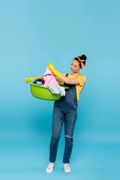 Full length view of housewife in denim overalls holding laundry bowl with clothes on blue — Stock Photo