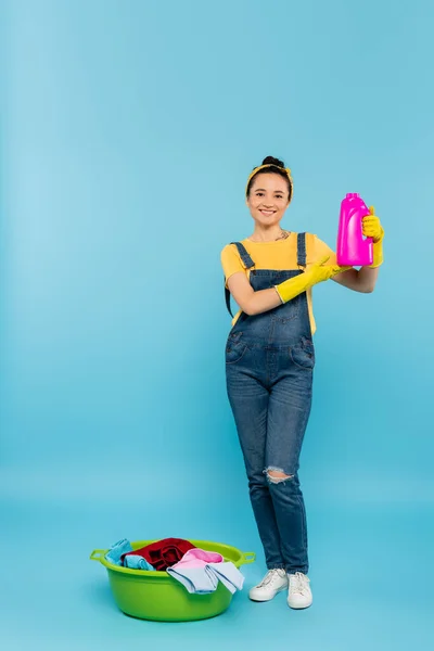 Heureuse femme au foyer en gants de caoutchouc jaune montrant bouteille de détergent près du bol à linge avec des vêtements sur bleu — Photo de stock