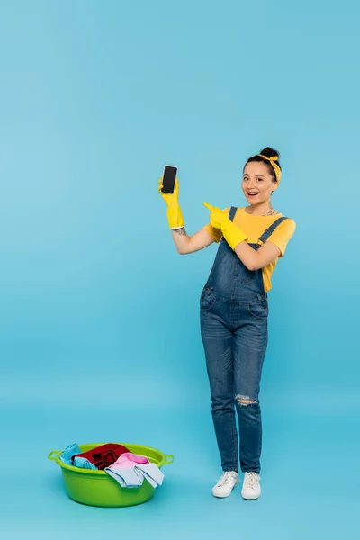 Femme au foyer souriante en salopette en denim pointant vers le smartphone près du bol à linge sur bleu — Photo de stock