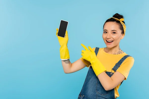 Mujer alegre en guantes de goma amarillos apuntando al teléfono inteligente con pantalla en blanco aislado en azul - foto de stock