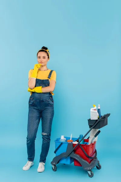 Cleaner in rubber gloves and denim overalls looking at camera near cart with cleaning supplies on blue — Stock Photo